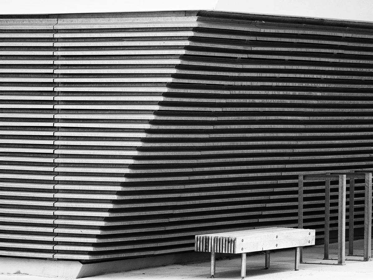 Fassade mit waagrechten Lamellen, davor eine schmale Bank – Schwarzweiß