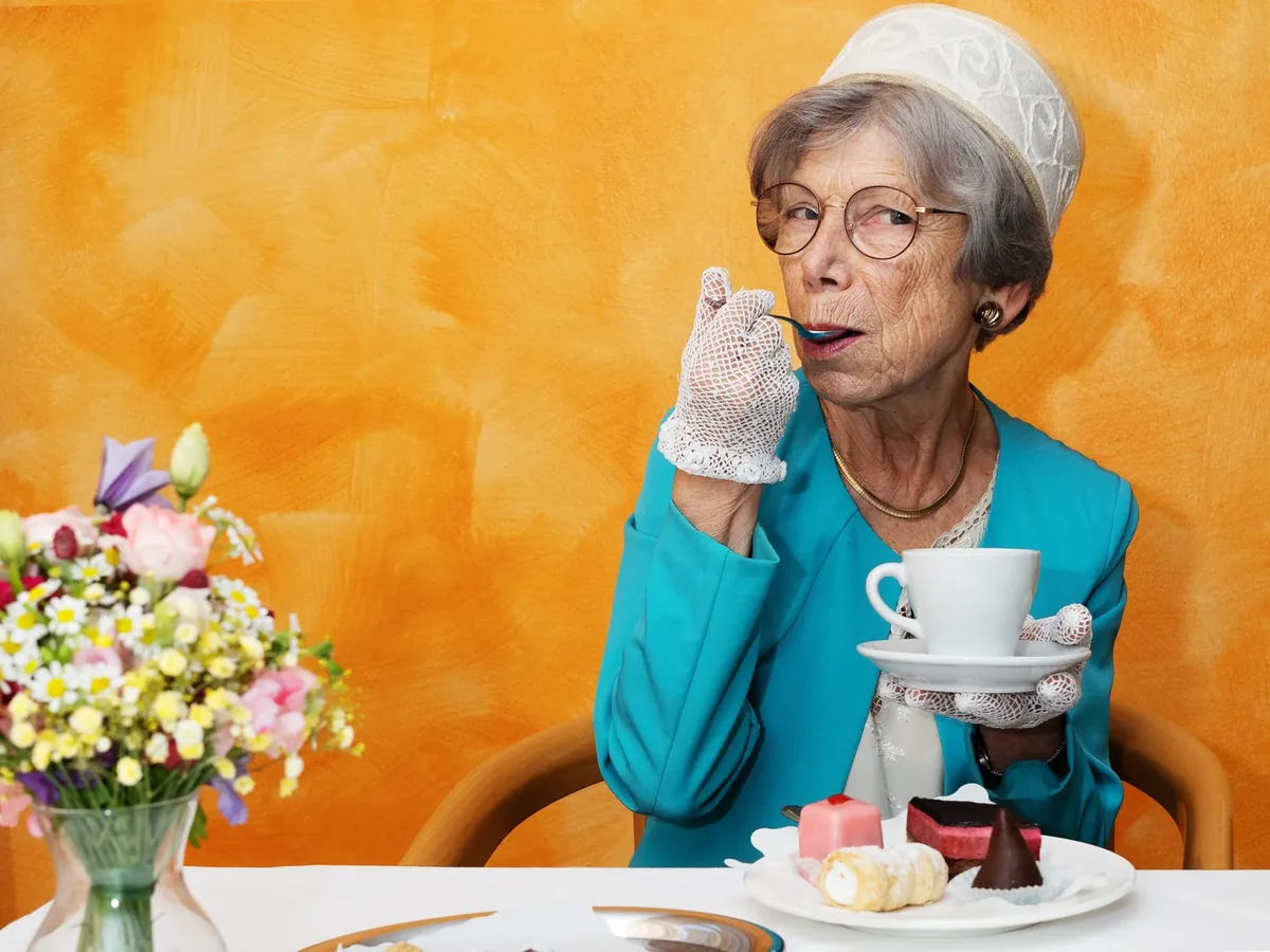 Ältere Dame mit Hut trinkt Kaffee vor orangem Hintergrund, daneben Blumen und Törtchen